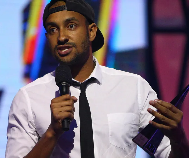 A man in a white shirt and tie holds a microphone and award on stage, wearing a cap backwards.