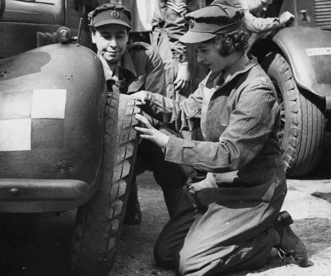 A woman in military uniform kneeling beside a vehicle, checking a tire with another uniformed person nearby.