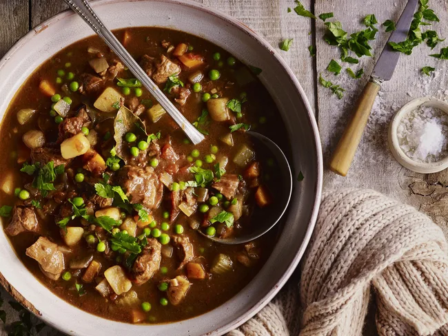 Hearty beef and vegetable stew in a bowl, garnished with parsley, with a cozy knitted cloth nearby.