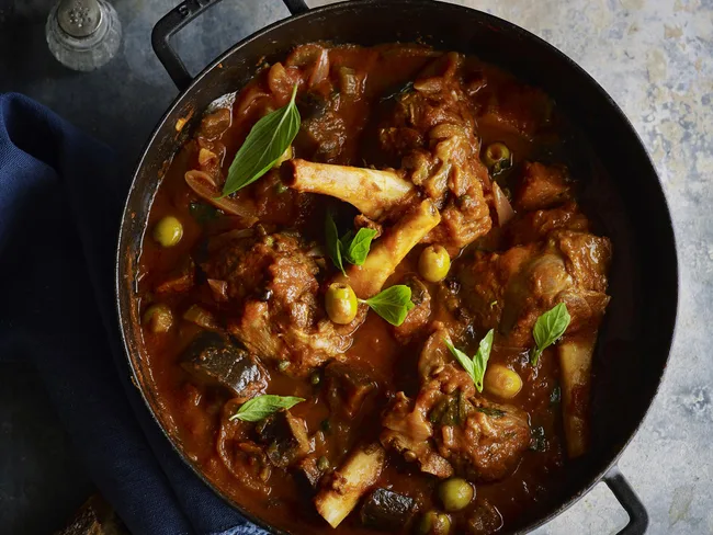 Slow-cooked lamb shanks in rich tomato sauce with herbs and olives in a black pot.