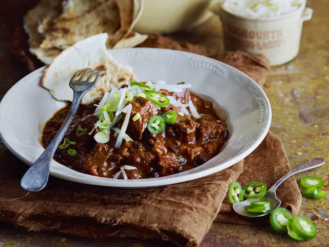 A bowl of chili with beef, topped with sliced green onions and jalape&ntilde;os, served with a fork and naan bread.