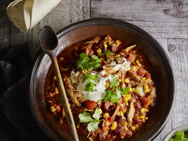 Bowl of chicken stew with corn, beans, tomatoes, topped with sour cream and cilantro, wooden spoon and tortillas on the side.