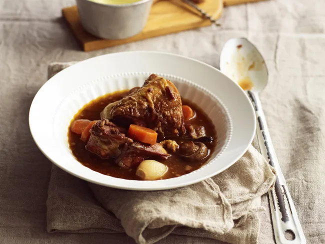 Slow-cooked beef stew with carrots in a white bowl, on a linen napkin with spoon and pot in the background.