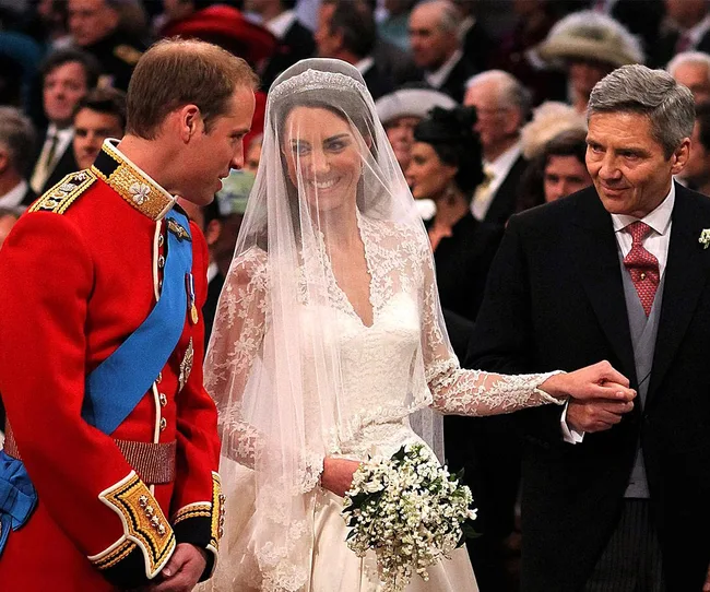 Prince in red uniform and bride in lace gown smiling at altar, holding bouquet, with older gray-haired man.
