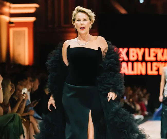 Woman in elegant black gown with puffy sleeves walking runway at fashion show, orange lit background.