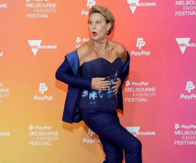 Person in a blue outfit poses dramatically at Melbourne Fashion Festival against a colorful background with logos.