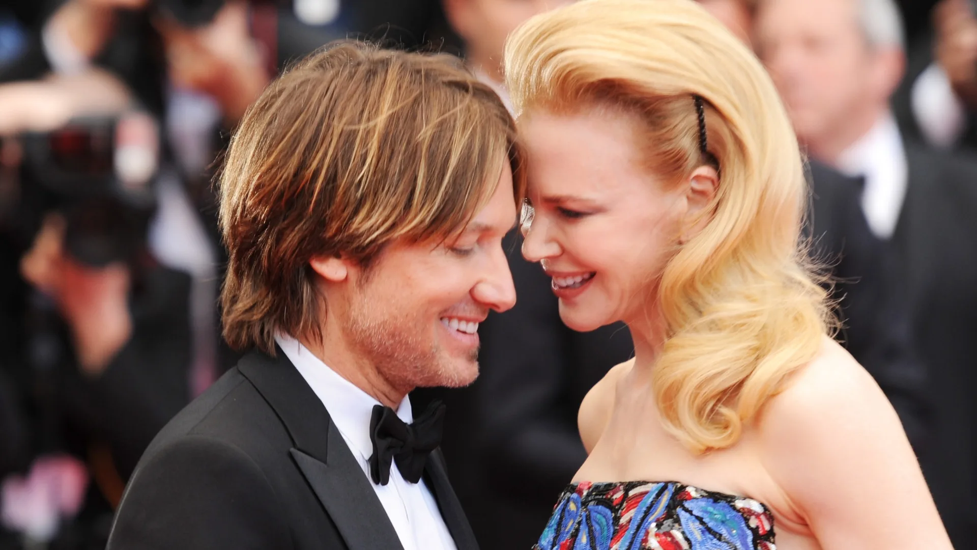 A couple smiles intimately on a red carpet, with cameras in the background.