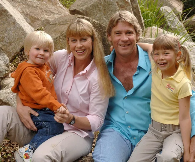 Family of four sitting outdoors, smiling; includes two adults and two children in a natural setting with rocks and grass.