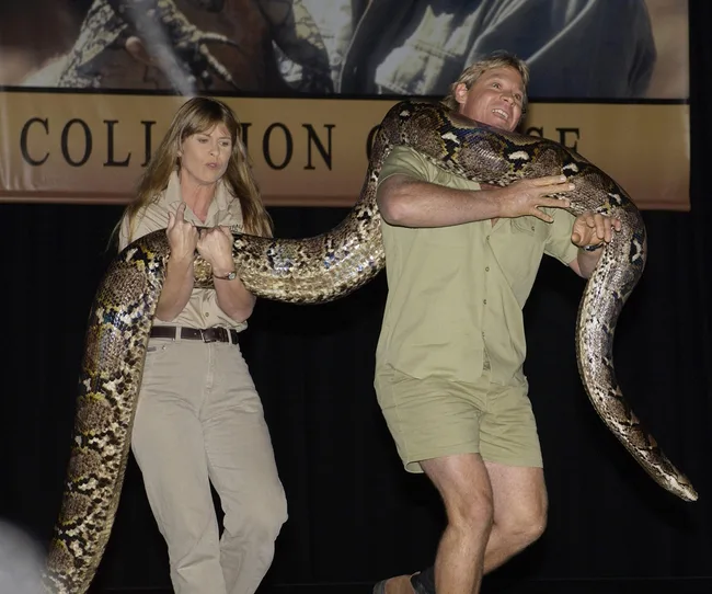 A man and woman in khaki outfits hold a large snake on stage during a wildlife demonstration.