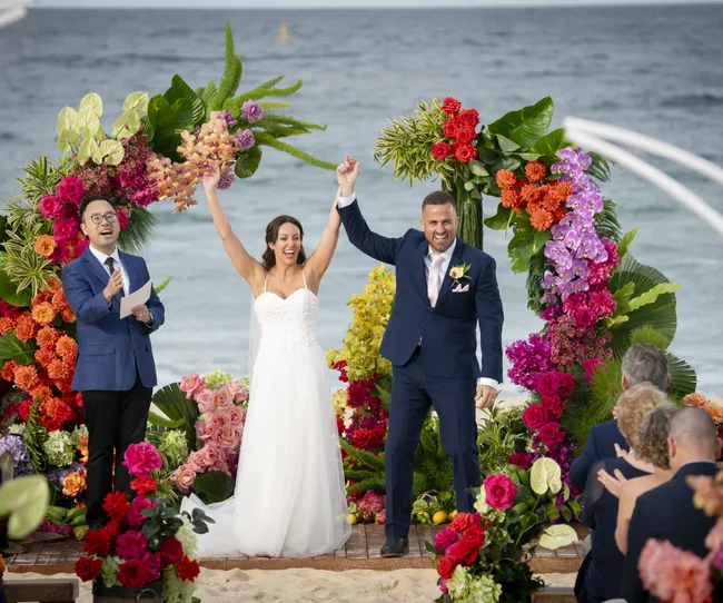 Bride and groom celebrate at a colorful beach wedding altar with an officiant, surrounded by vibrant flowers.