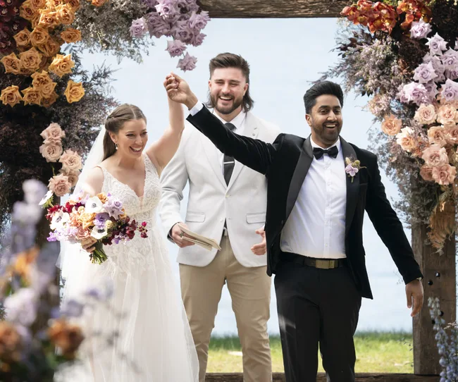 Bride and groom celebrating under a floral arch with officiant, smiling and holding hands.