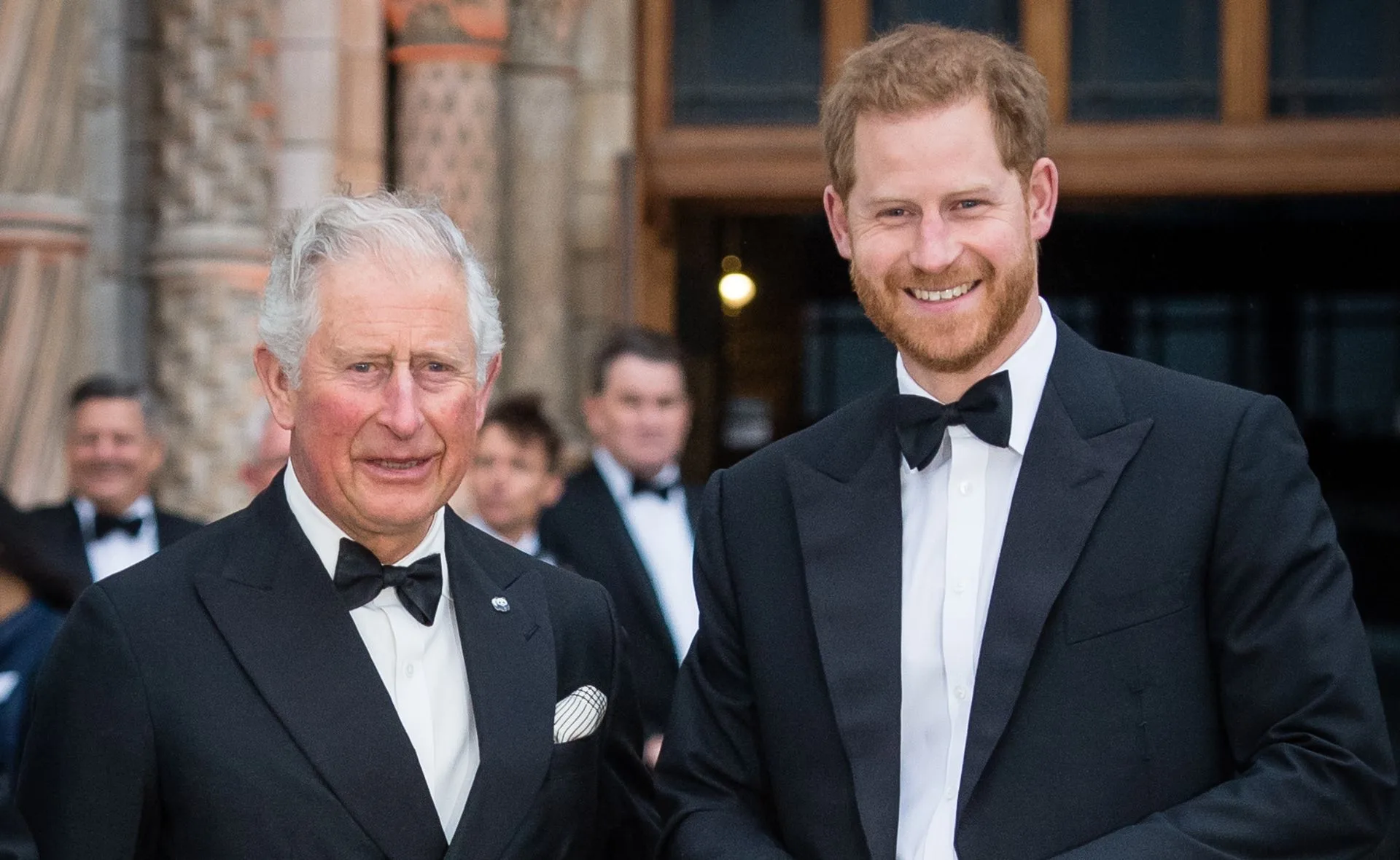 Two men in tuxedos smiling, standing outside a building with ornate stonework, surrounded by people in formal attire.