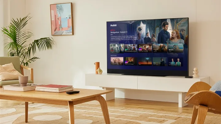 Living room with TV showing "Bridgerton: Season 3" on Netflix, wooden table and sofa, framed art on wall.