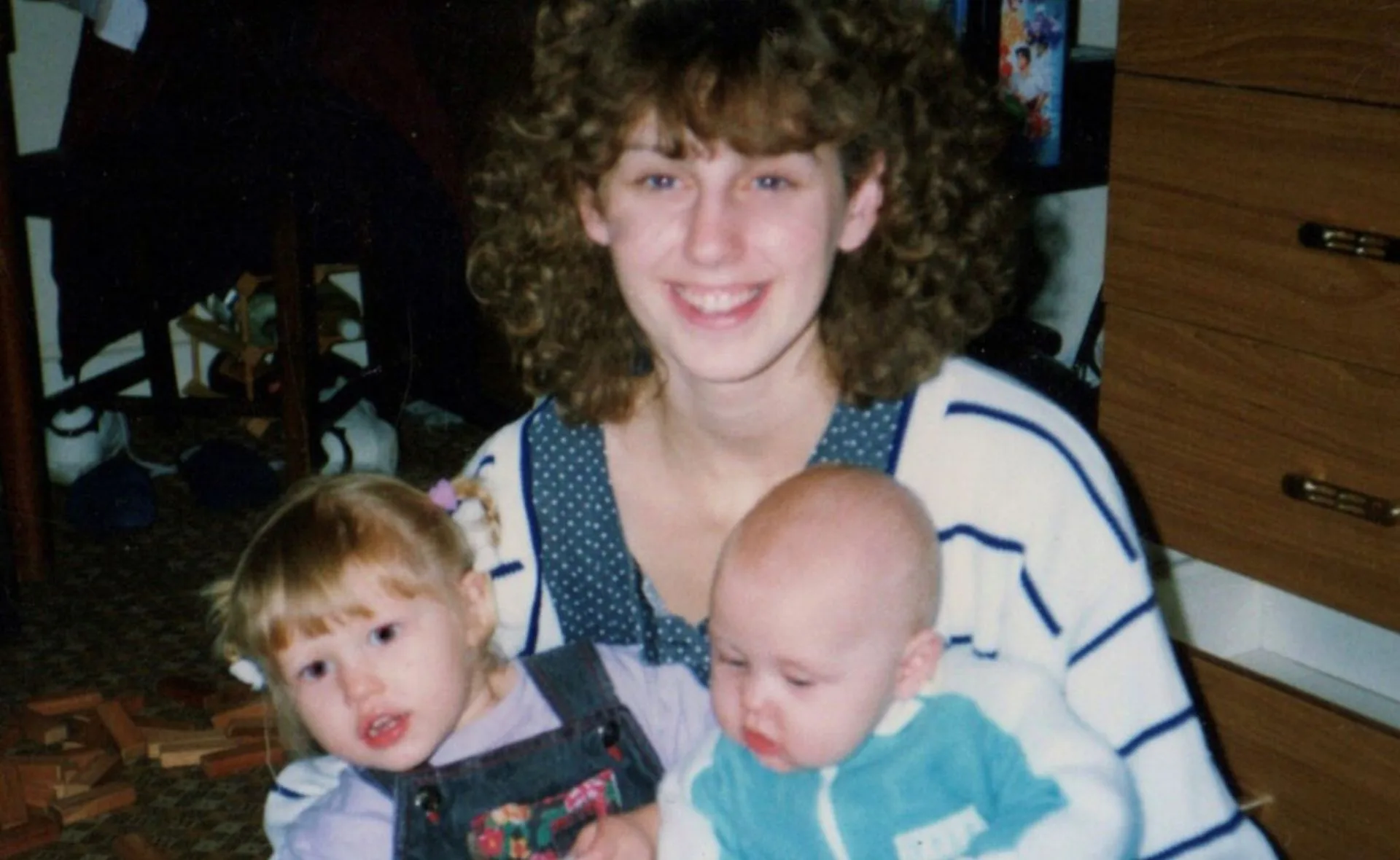 Young woman with curly hair smiles while holding a toddler and a baby indoors.