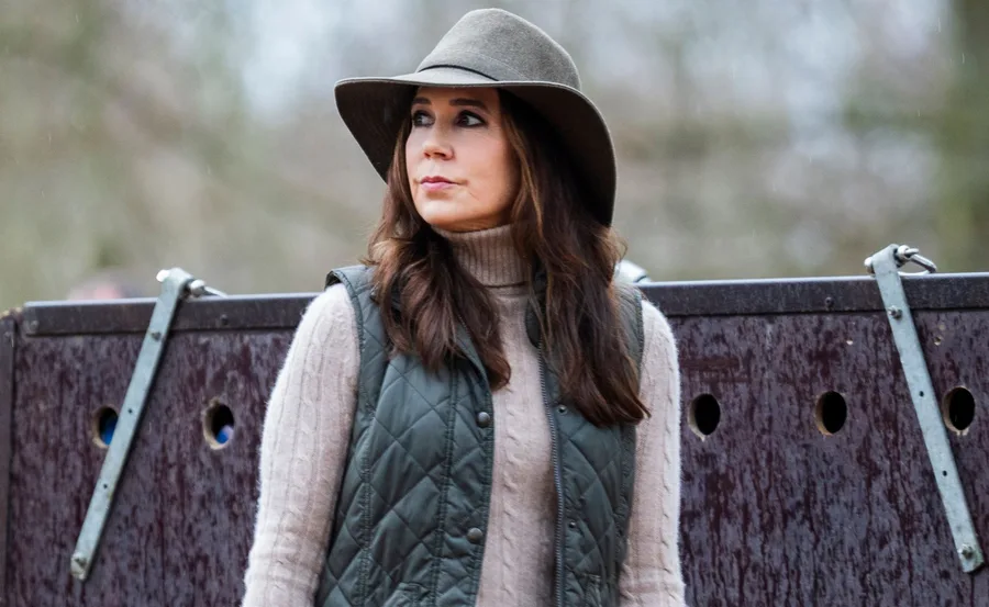Woman in hat and quilted vest outdoors, standing near wooden panel, looking off to the side.