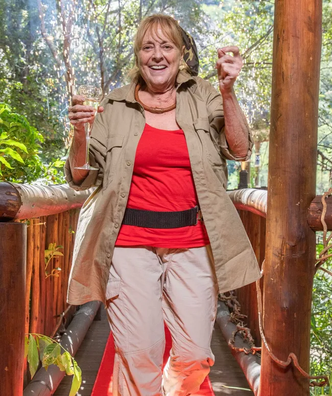 A woman in a red top and khaki clothes holding a champagne glass, smiling, standing on a wooden bridge in a forest.