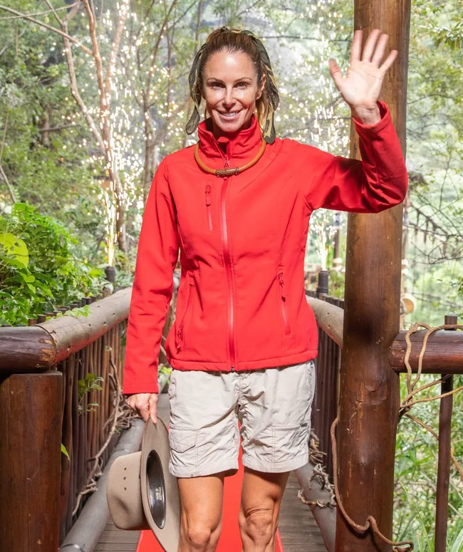 Woman in red jacket and beige shorts waving, holding a hat, standing on a jungle bridge with sparklers in the background.