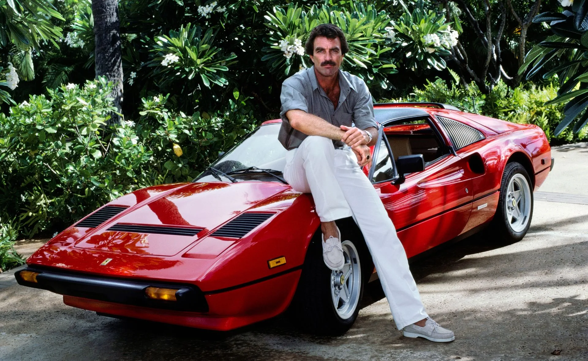 Tom Selleck sits on red car for a Magnum PI shoot. He's wearing white pants and a Hawaiian button-up shirt.