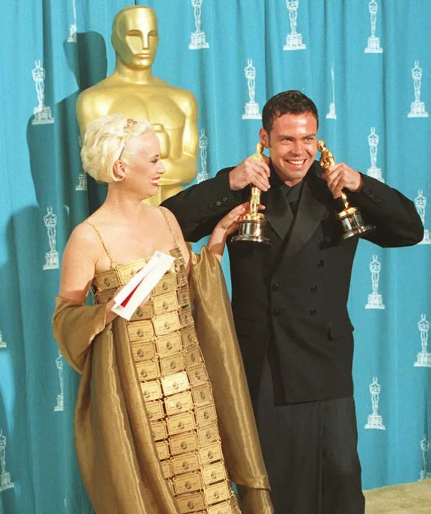 Tim and Lizzy smile at the Oscar in 1995. Tim holds two Oscars by his face, cheesing big. Lizzy wears a dress made from gold credit cards.