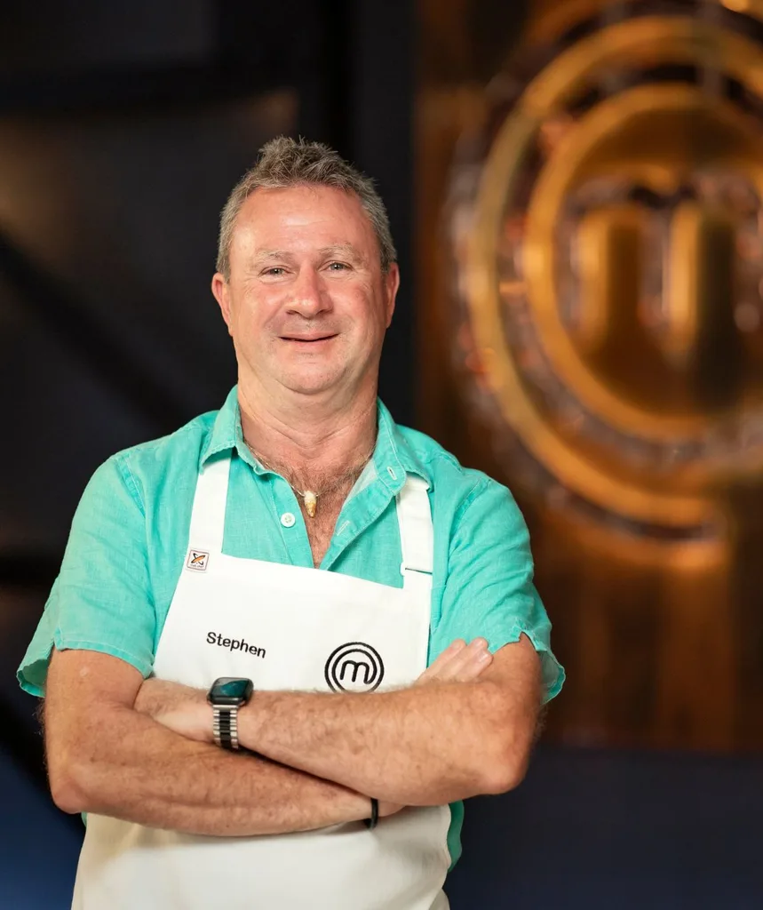 MasterChef favourite Steve Dennis smiles, wearing crocodile-tooth necklace, blue shirt and white apron with arms crossed in front of MasterChef logo