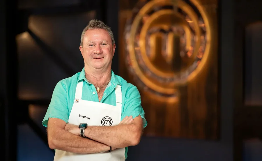 MasterChef Stephen Dennis crosses arms in front of MasterChef logo wearing white apron and blue collared shirt
