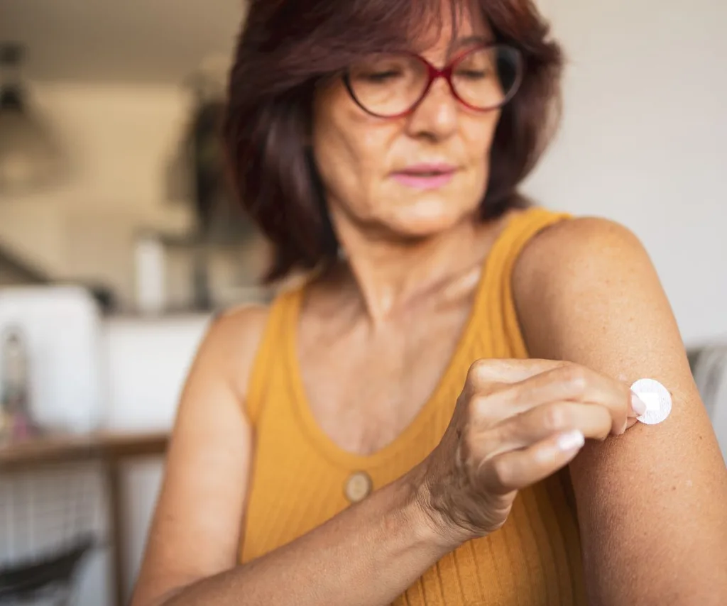 Woman in her 50s wearing orange tank top and red-rimmed glasses places patch on her upper arm