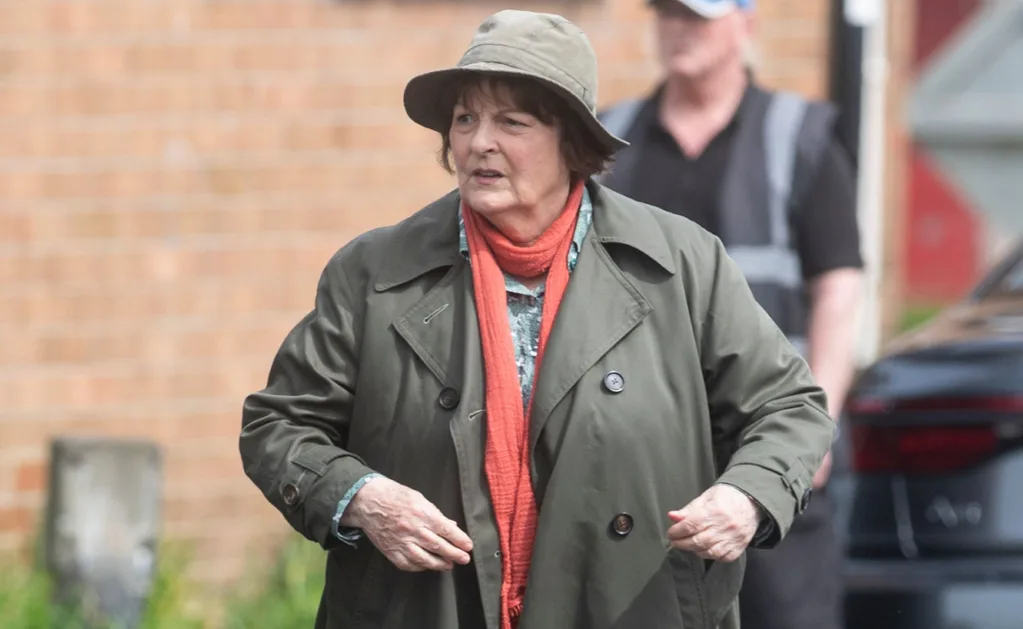 Brenda Blethyn as Vera is standing looking to the side, wearing a trench, hat and thin red/orange scarf while filming Vera season finale