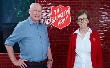 Ray Meagher and Salvos Chief Miriam Gluyas stand in front of red wall with The Salvation Army emblem between them