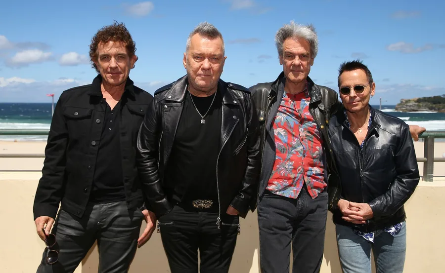 Cold Chisel band stands outside in black jackets against a beach backdrop