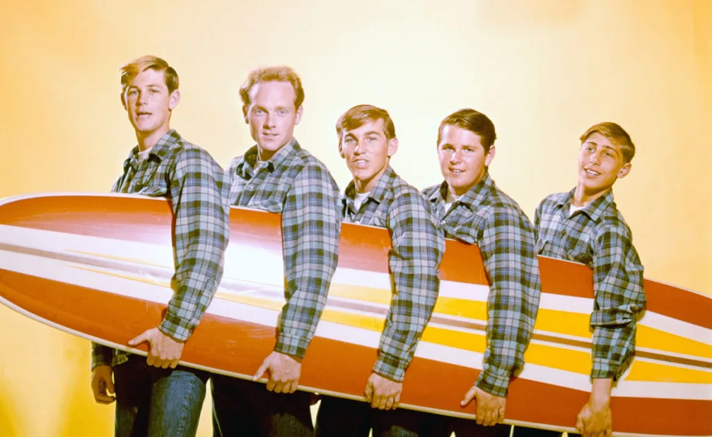 The Beach Boys posing in flannel shirts, holding a retro surfboard