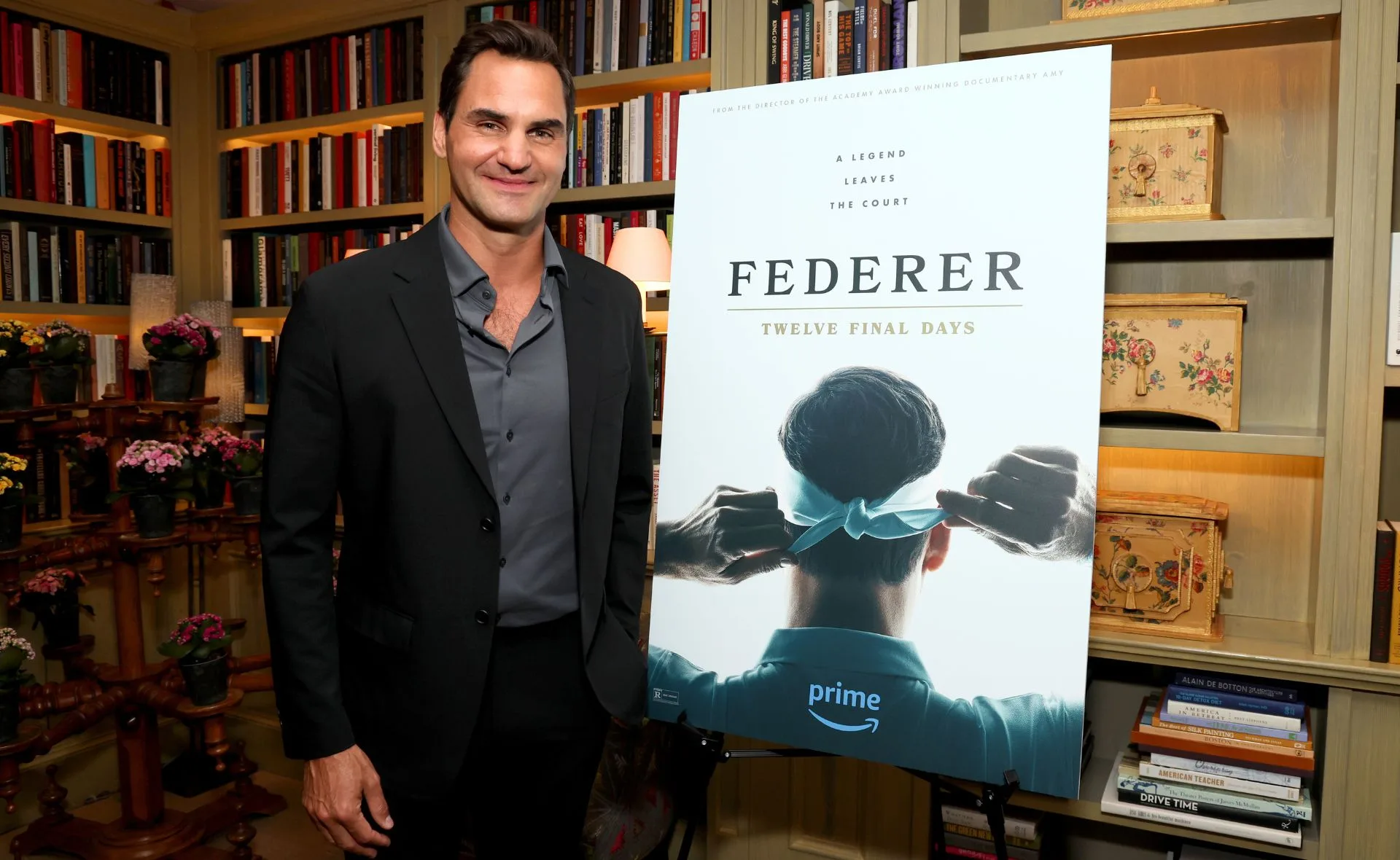 Man standing next to a poster titled "Federer: Twelve Final Days" inside a room with bookshelves.