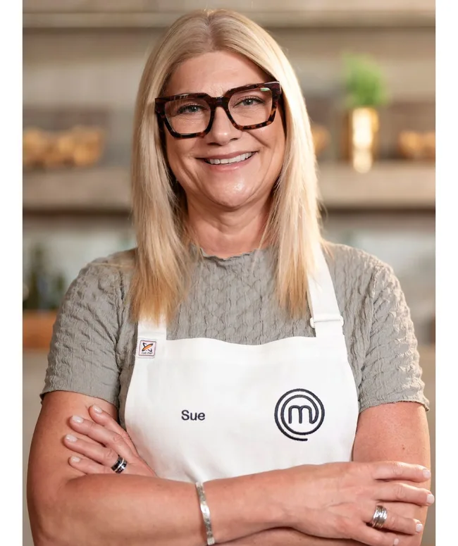Woman with long blonde hair wearing glasses and a "MasterChef" apron, smiles with arms crossed.