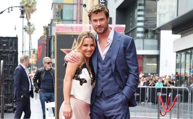 Elsa wearing a white dress with black lace trim stands with husband Chris Hemsworth, wearing blue suit with vest, smiling