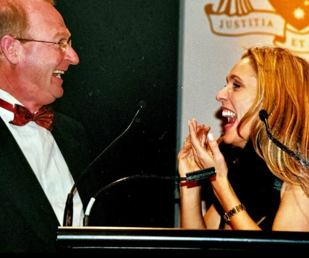 Tottie Goldsmith beams up at John Blackman, both wearing black, standing in front of a podium
