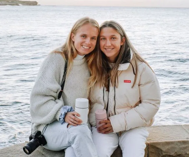 Two people smiling, sitting by the sea holding thermos cups, with a camera hanging from one person's shoulder.
