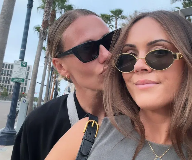 Two people wearing sunglasses, embracing and smiling, outdoors with palm trees and city buildings in the background.