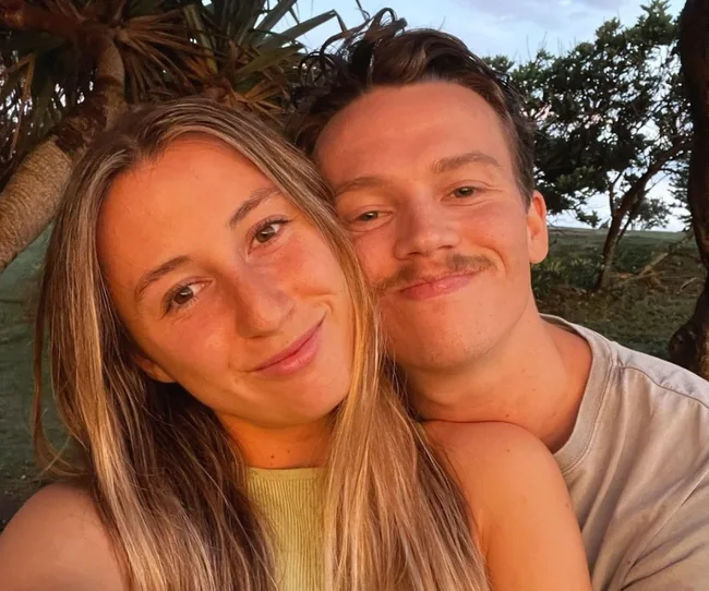 A smiling couple takes a close-up selfie outdoors near trees during sunset.