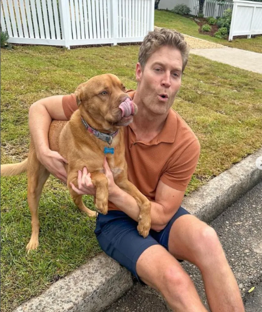 Dr Chris in an orange shirt holding a dog that's licking its lips 