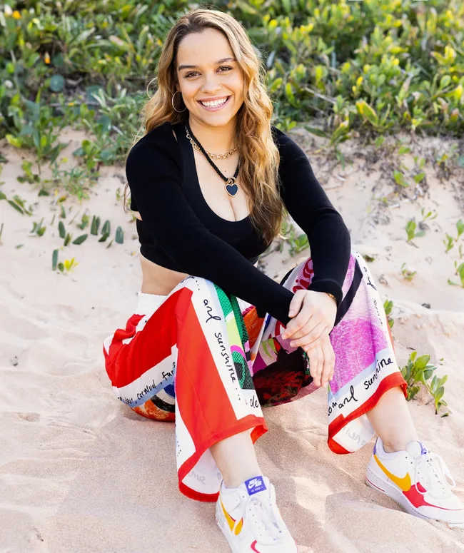Young woman sitting on sandy ground, wearing colorful pants and a black top, smiling with greenery in the background.