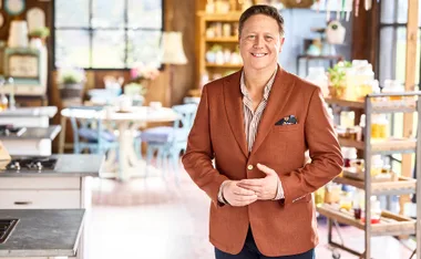 Man in a brown blazer smiling in a cozy, well-lit kitchen setting.