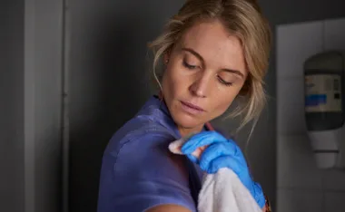 A woman in scrubs and gloves wipes her arm with a cloth, appearing focused.