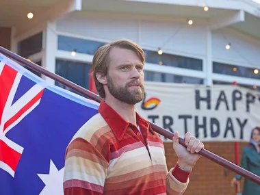 A bearded Jesse Spencer, playing Tony in Last Days Of The Space Age, carrying an Australian flag on his shoulder.