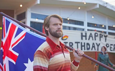 A bearded Jesse Spencer, playing Tony in Last Days Of The Space Age, carrying an Australian flag on his shoulder.