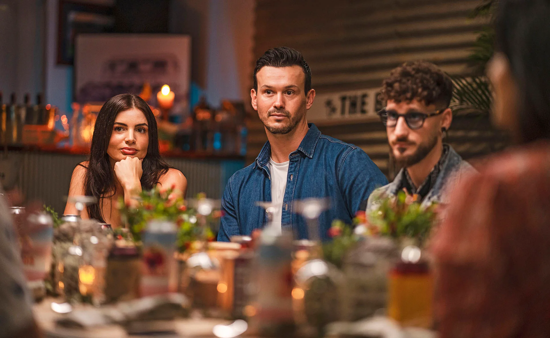 Three people sit at a dinner table, with plants and candles around, in a warmly lit setting.