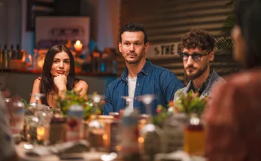 Three people sit at a dinner table, with plants and candles around, in a warmly lit setting.
