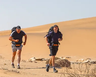 Billy and Oscar Brownless, with backpacks on, are running through sand dunes on The Amazing Race Australia: Celebrity Edition.