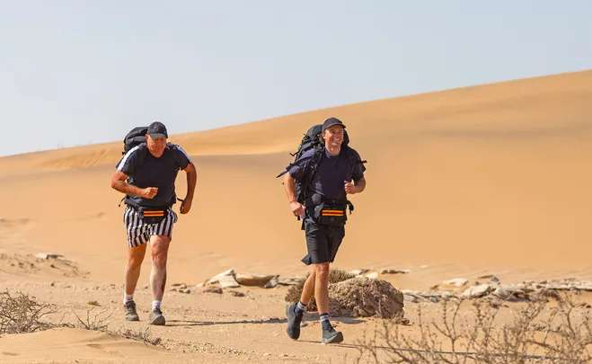 Billy and Oscar Brownless, with backpacks on, are running through sand dunes on The Amazing Race Australia: Celebrity Edition.