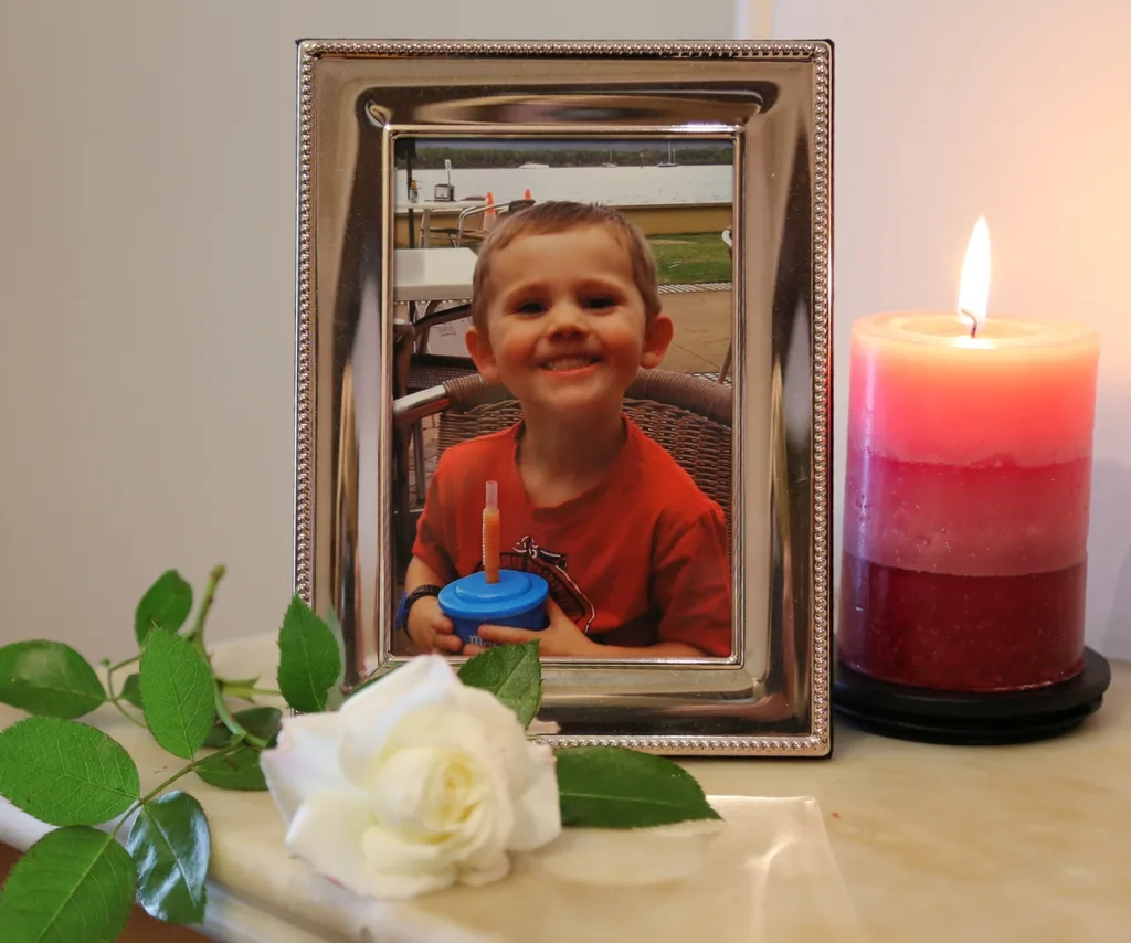 A picture frame with an image of a smiling William Tyrrell, a white flower and a lit candle.