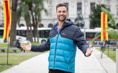 Man in a blue puffer jacket smiling with arms outstretched in a city park setting, flags in the background.