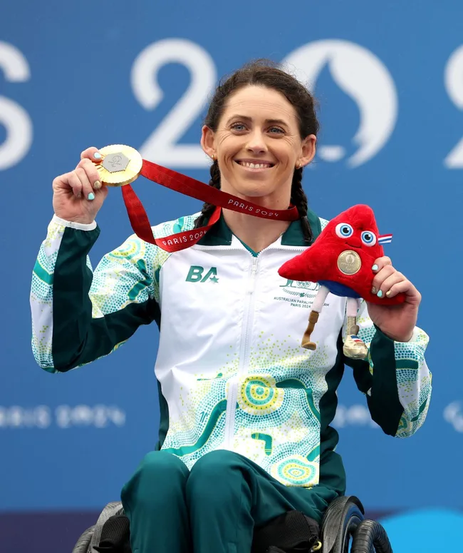 Australian Paralympian in wheelchair holding a gold medal and red plush mascot, smiling at the Paris 2024 event.